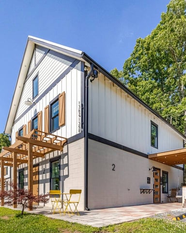 Modern farmhouse with white and gray exterior, wooden pergolas, patio seating, surrounded by trees and green lawn