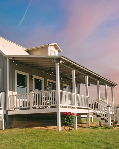 Modern farmhouse with metal roof, white railings, and wraparound porch at sunset overlooking green fields