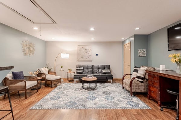 Finished basement living room with black leather sofa, wooden armchairs, patterned area rug, and wood furniture on hardwood flooring.