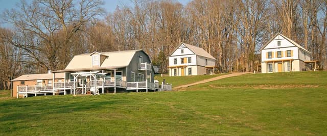 Three farmhouse-style buildings with white trim and metal roofs on expansive green lawn surrounded by bare trees
