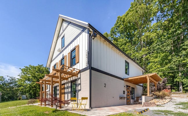 Modern farmhouse with white and gray exterior, wooden pergolas, patio seating, surrounded by trees and green lawn